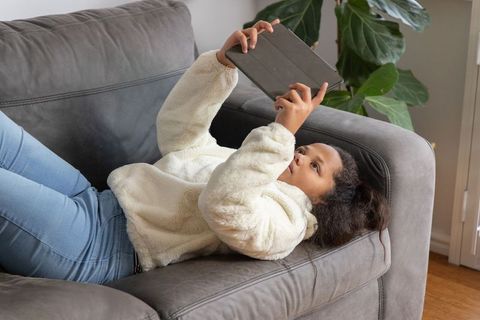 Young girl reclining on gray sofa using tablet in cozy fleece and jeans