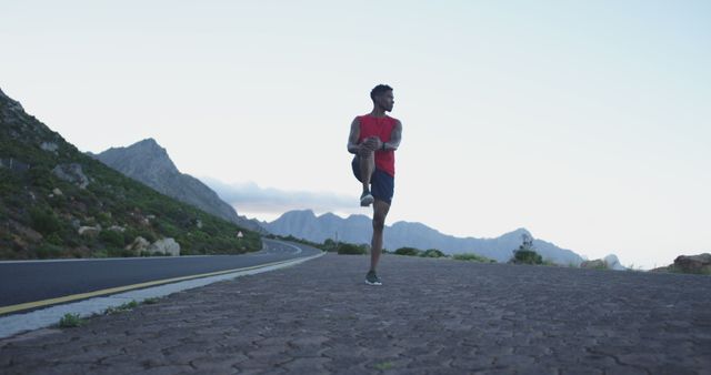 Man is stretching on a mountain road during morning, with a clear sky and mountains in the background. Useful for promoting healthy lifestyles, fitness programs, outdoor activities, or exercise apparel.