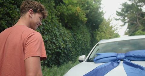 Young man receiving new white car with oversized blue bow on suburban driveway
