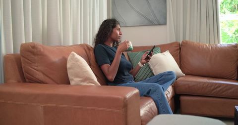 African american woman relaxing on tan leather sectional holding mug and using smartphone