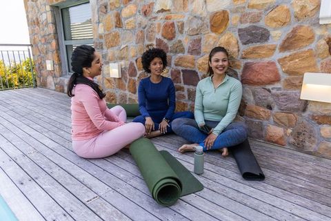 Diverse women chatting on wooden terrace after outdoor yoga by stone wall, wellness break
