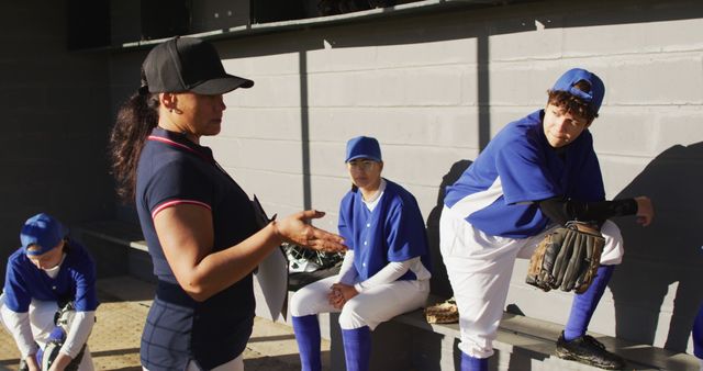 The image shows a youth baseball team in uniforms listening to a coach, likely discussing game strategy or providing instructions during a break. Ideal for illustrating themes of teamwork, sportsmanship, youth athletics, and training sessions. Can be used for school sports programs, coaching tutorials, and team-building materials.
