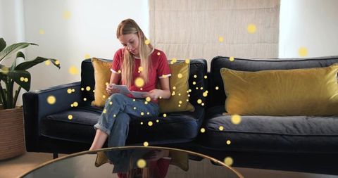 Young woman writing diary on dark velvet sofa with mustard pillows in cozy living room