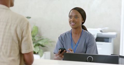 African american medical receptionist greeting patient at clinic desk with smartphone