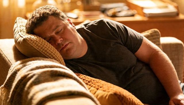 Middle-aged man napping on cozy beige sofa with textured throws and warm lamp light