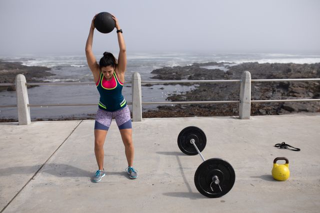 Front view of a strong Caucasian woman with long dark hair wearing sportswear exercising outdoors by the seaside on a sunny day, strength training lifting ball, rubber tape, kettlebell and barbells next to her.