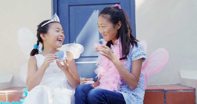 Two Asian girls are enjoying a playful tea party outdoors, dressed in whimsical costumes with fairy wings and a tiara. Their laughter and imaginative play create a joyful atmosphere, celebrating the innocence and creativity of childhood.