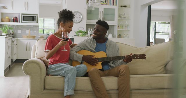 Image of happy african american couple sitting on sofa, singing and playing guitar. love, relationship, togetherness and spending quality time at home.