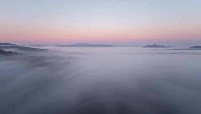 Aerial pastel sunrise over fog-filled valley with flowing mist and distant hills