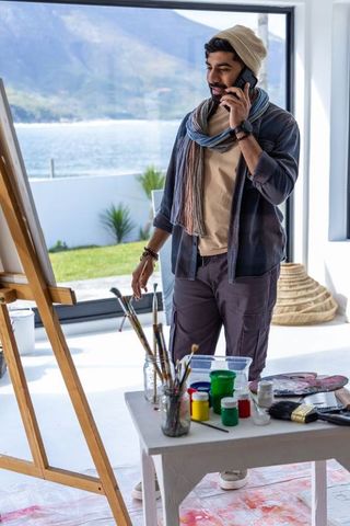 Bearded male artist phoning while painting in bright coastal studio with easel and palette