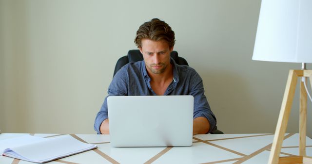 Young man is working intensely on a laptop in a modern, minimalistic office. Image portrays professionalism, dedication, and focus, suggesting uses in business blogs, tech articles, and productivity content.