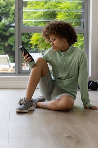 African american woman resting by window after home workout checking phone for progress