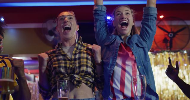 Young women enthusiastically cheering while watching a sports game at a bar, capturing the excitement and camaraderie of a night out with friends. Perfect for advertisements focusing on social gatherings, sports events, lifestyle blogs, and promotional materials for bars and entertainment venues.
