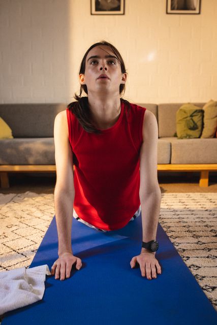 Non-binary person practicing yoga on a blue mat in a cozy living room. Ideal for articles or advertisements focusing on home fitness, gender fluidity, mindfulness, and self-care routines.