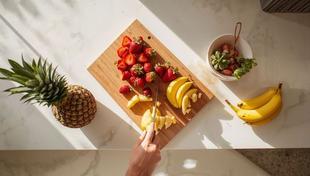 Hand slicing banana on wooden cutting board with strawberries and pineapple, sunlit kitchen