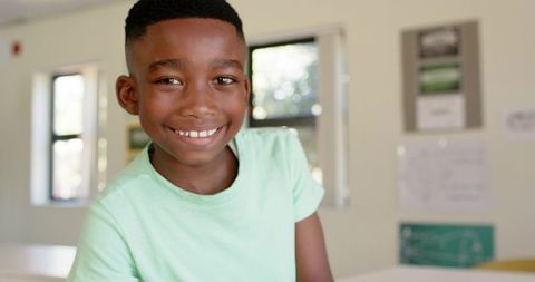 African american boy smiling in sunlit classroom leaning on desk, happy school portrait