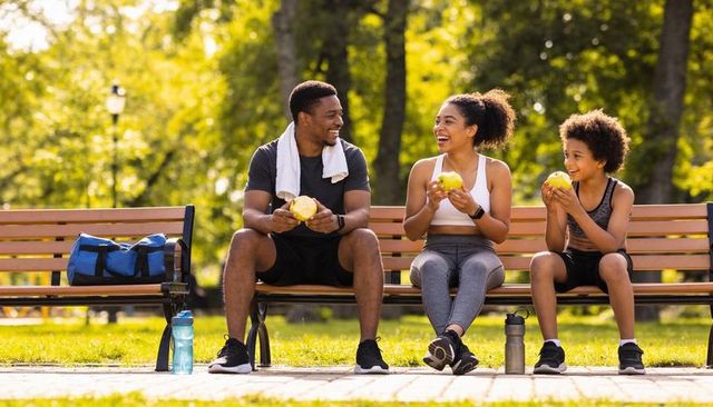 Family laughing and eating apples on park bench after workout, hydration and fitness