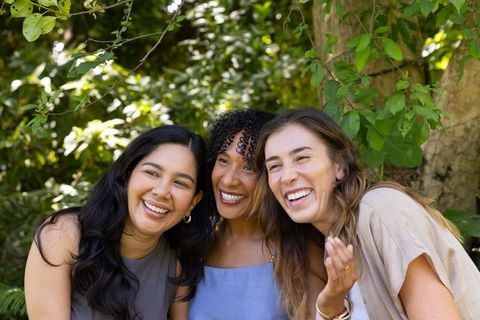Diverse female friends laughing together outdoors under green canopy, candid summer trio