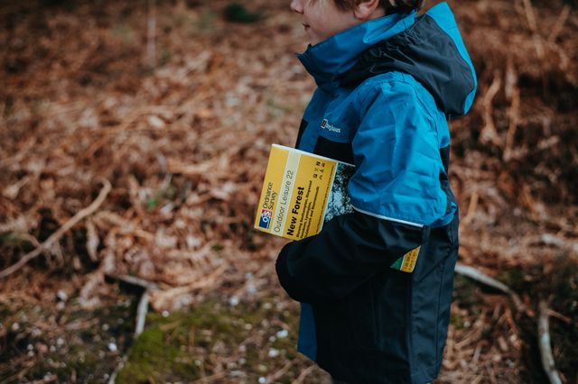 A child wearing a blue and black jacket is hiking in a forest. They are holding a map. Ideal for portraying family vacations, outdoor activities, youth exploration, and adventure travel guides.
