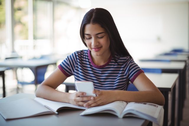 This image depicts a schoolgirl using a mobile phone while studying in a classroom. It can be used to illustrate themes related to modern education, the integration of technology in learning, student life, and academic focus. Ideal for educational websites, blogs about student life, and articles on the impact of technology in education.