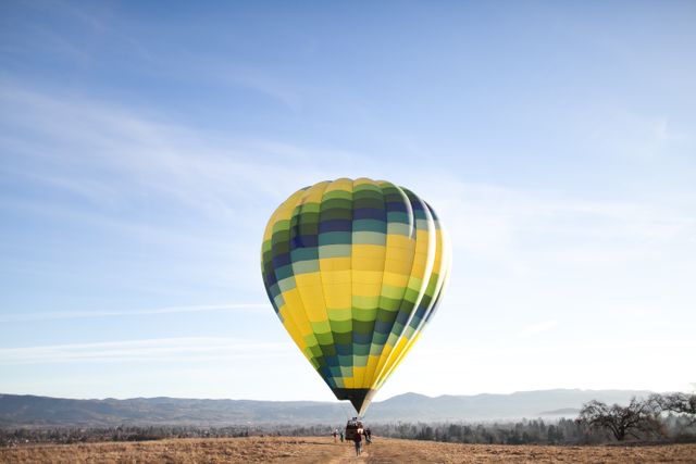 Hot air balloon featuring vibrant yellow, green and blue colors preparing for takeoff in open field with clear blue sky and distant mountains. Suitable for travel and adventure themes, promoting outdoor activities, or illustrating peaceful scenic landscapes.