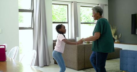 African grandmother and granddaughter dancing in sunlit living room, joyful family bond
