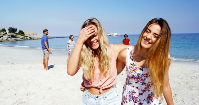 Group of friends enjoying time at beach on sunny day. Main focus on two young women at front smiling and walking together. Perfect for themes of friendship, summer vacation, beach activities, and relaxation in nature.