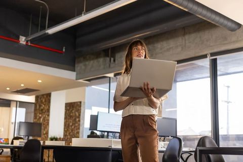 Woman standing holding laptop and looking upward in modern open-plan office with daylight