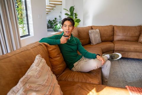 Asian man relaxing on brown leather sofa using voice remote in modern sunlit living room