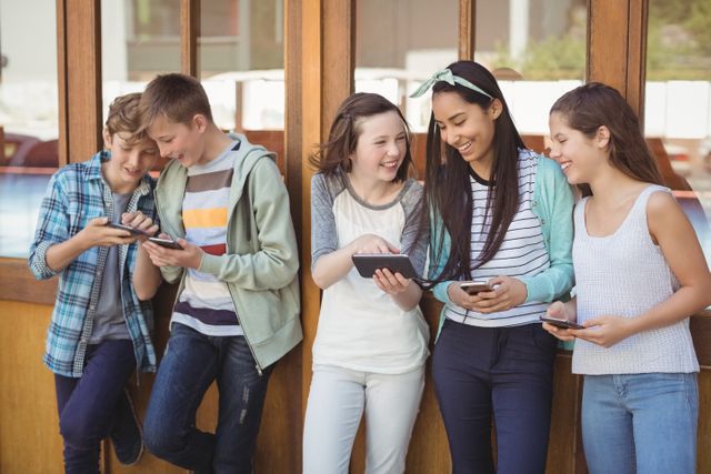 Group of diverse school friends standing in corridor, using mobile phones and smiling. Ideal for themes related to technology, social media, youth culture, education, and friendship. Can be used in educational materials, social media campaigns, and advertisements targeting teenagers.