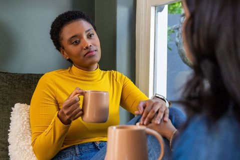 Sharing cozy coffee and conversation on sofa by window, diverse friends holding mugs