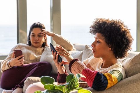 Sunlit diverse friends relaxing and browsing devices on sofa with seaside view