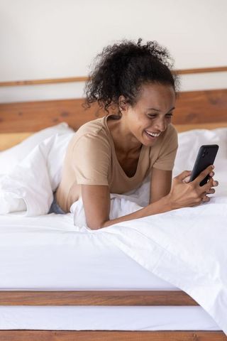 African american woman smiling and using smartphone while lying on bed with white bedding