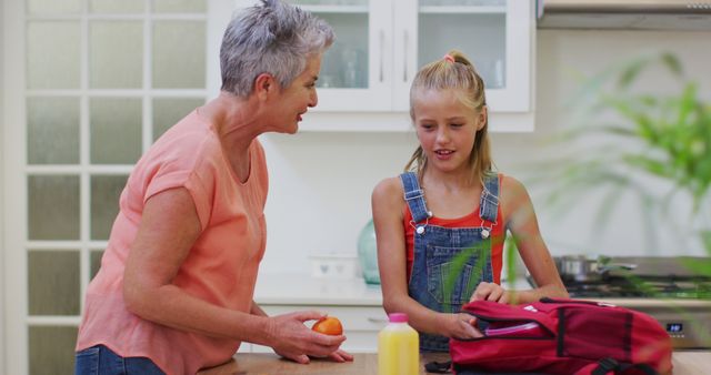 Caucasian grandmother in kitchen preparing packed lunch talking with granddaughter giving her fruit. family spending time together at home.