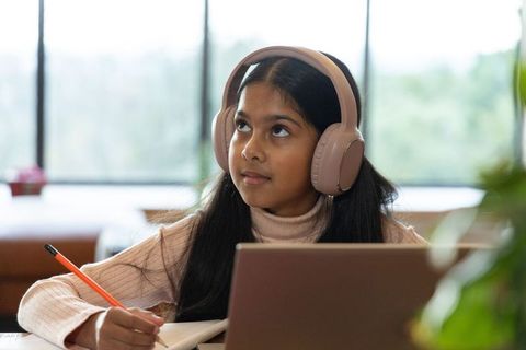 Indian preteen girl wearing headphones studying at laptop, writing notes in home study