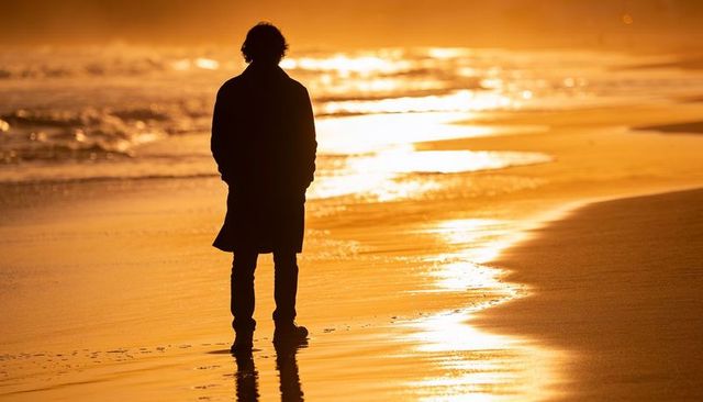Standing silhouette on golden shoreline at sunset with reflective waves and footprints