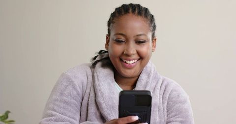 Smiling african american woman wearing plush bathrobe using smartphone for morning messages