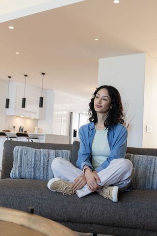 Woman sitting cross-legged on sofa in modern minimalist living room, looking thoughtful