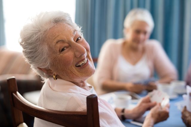 Senior woman enjoying a game of cards with friends in a nursing home. Ideal for use in materials promoting senior living communities, elderly care, social activities for seniors, and retirement lifestyle. Highlights themes of friendship, happiness, and active aging.