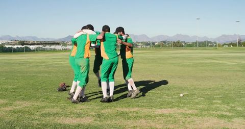 Cricket team huddling on sunlit outfield wearing green orange uniforms showing teamwork