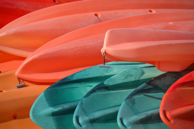Kayaks of various colors are stacked outdoors under bright sunlight. Vibrant hues of orange, green, and red highlight the kayaks' sleek lines. Perfect for promoting water sports, recreational activities, outdoor adventures, and summer excursions. Suitable for use in travel brochures, sport equipment advertisements, and adventure tour company websites.