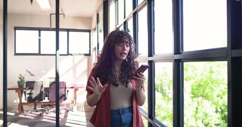 Young woman conducting video call while standing by sunlit glass wall in modern office