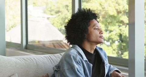 African American man gazing out window, relaxing on sofa, wearing denim jacket and earbud