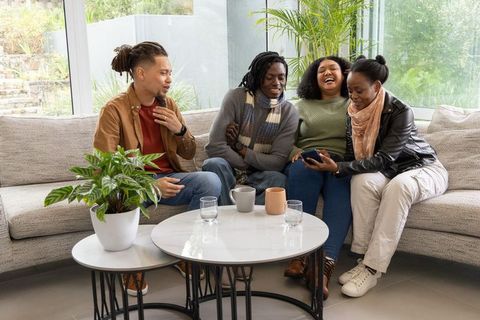 African american friends laughing and sharing smartphone on sofa in bright living room