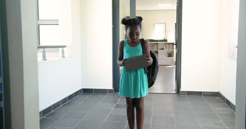 African american girl using tablet in school corridor wearing teal dress and backpack