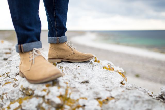 A close-up of a person wearing suede boots, standing on rugged coastal rocks with the ocean in the background. This scene suggests outdoor exploration and adventure. It is perfect for use in travel blogs, outdoor fashion campaigns, or articles about tourism and nature.
