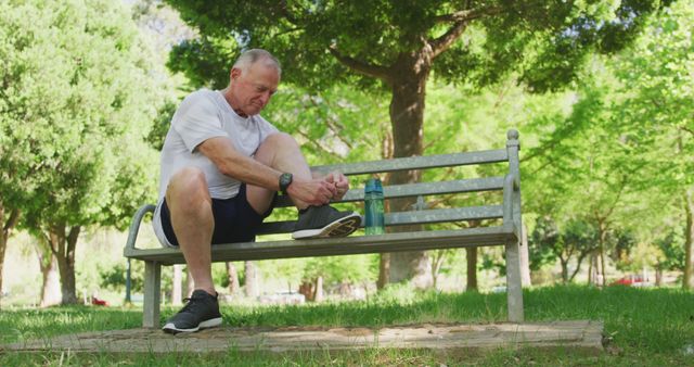 Senior man is sitting on park bench tying shoelaces while preparing for exercise. Water bottle indicating hydration rests beside him. Scene showcasing an active and healthy lifestyle for mature adults. Ideal for promoting fitness, outdoor activities, and healthy aging.