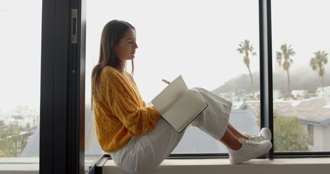 Woman writing in notebook by window overlooking cityscape