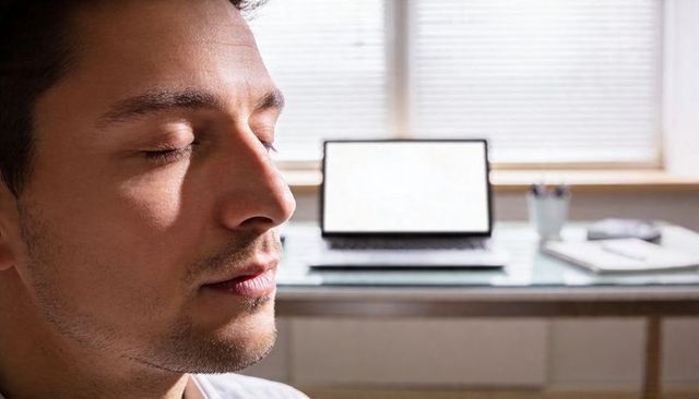 Sunlit close-up of man practicing mindfulness at home desk, closing eyes in peaceful pause