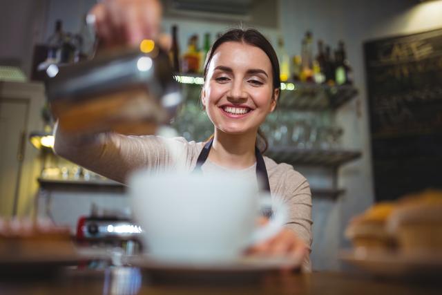Waitress smiling while making a cup of coffee at a cafe counter. Ideal for use in articles or advertisements related to coffee shops, customer service, hospitality industry, and barista training. Can also be used for promoting cafes and coffee-related products.